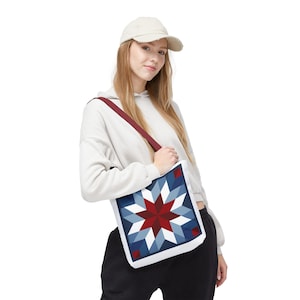 May include: A white tote bag with a geometric star pattern in shades of red, white, and blue. The bag has a burgundy strap and is carried over the shoulder. The bag is a square shape and has a white border.