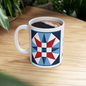 May include: White ceramic coffee mug with a geometric star design in red, white, and blue. The mug is filled with dark coffee and sits on a wooden surface. The mug has a white handle.