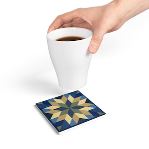 May include: A white ceramic mug filled with a dark liquid is held above a square coaster. The coaster features a geometric starburst pattern in shades of blue, tan, and green, with a dark blue border.
