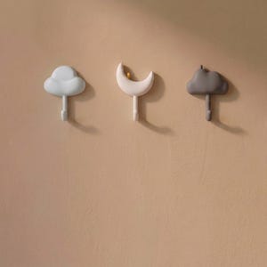 May include: Three wall hooks featuring celestial and weather-themed designs. The left hook is a light blue cloud, the center hook is a pale pink crescent moon, and the right hook is a dark gray cloud. Each piece includes a small downward-facing hook.