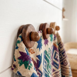 May include: Three circular dark brown wooden wall hooks mounted on a white surface. A woven textile with a multi-colored geometric pattern in shades of purple, green, blue, and orange is draped over the hooks, hanging down in folds.