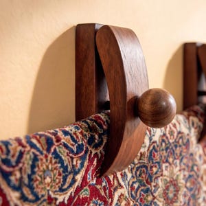 May include: A dark brown wooden wall-mounted clamp featuring a curved arm and a spherical knob, securely holding a section of a woven textile with a detailed red, blue, and cream-colored intricate floral pattern across its surface.