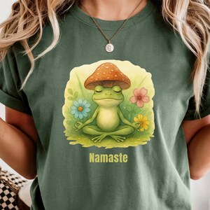 May include: A moss green t-shirt featuring a graphic of a green frog sitting in a lotus position with a mushroom cap on its head, surrounded by colorful flowers. The word Namaste is printed in yellow text below the frog illustration.