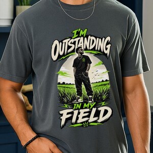 May include: A charcoal grey short-sleeve t-shirt featuring a graphic print of a person standing in a field. The text "I'M OUTSTANDING IN MY FIELD" is printed in white and lime green lettering above and below the central illustration.