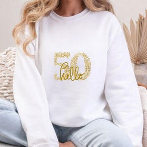 May include: White sweatshirt with a gold glitter design. The design features the number "50" and the word "hello" in a script font. The sweatshirt is a casual style, suitable for a birthday celebration.