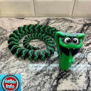 May include: A green and black articulated snake toy with a cartoonish design. The snake's body is black with green zigzag patterns, and the head is green with large eyes and a forked tongue. The toy is coiled on a speckled surface.