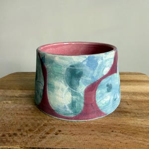 May include: A cylindrical ceramic planter with a wide opening. The interior is a deep pink, while the exterior features a pattern of blue and teal circles with maroon accents. The planter sits on a wooden surface.