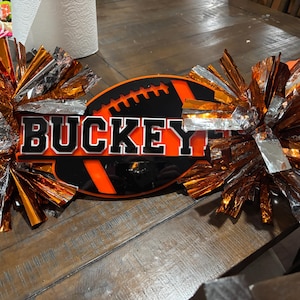 May include: A decorative sign featuring the word BUCKEYES in black lettering with a white outline, layered over an orange and black football graphic. The sign is flanked by two large, metallic orange and silver cheerleading pom-poms.