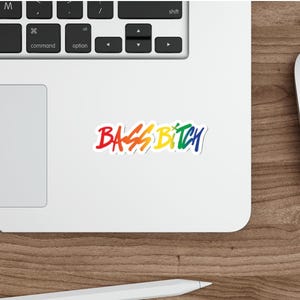 May include: A colorful sticker with the words "BASS BITCH" in a rainbow gradient. The sticker is on a white surface, next to a laptop keyboard, a computer mouse, and a stylus on a wooden surface.