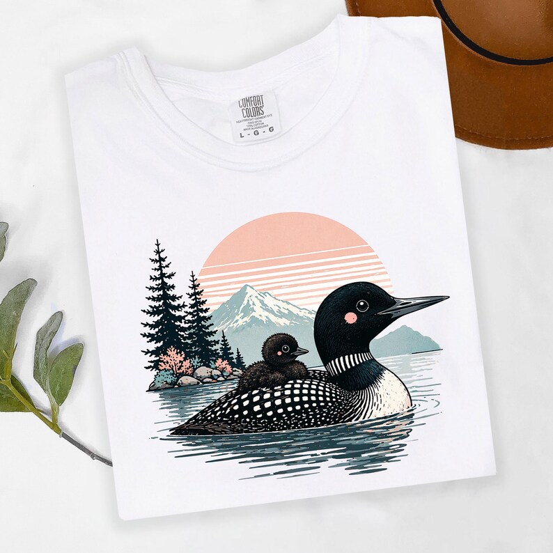 以下が含まれることがあります： 湖にいるアビとヒナのグラフィックが描かれた白いTシャツ。ピンクとオレンジの夕日、山、木々が背景に描かれています。「Comfort Colors」とラベルされ、サイズはL-G-Oです。