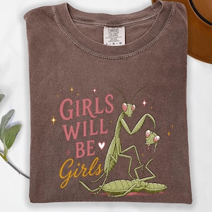 May include: Brown t-shirt with the text "GIRLS WILL BE Girls" in pink and yellow. The shirt features a graphic of green praying mantises. The shirt is made by Comfort Colors.