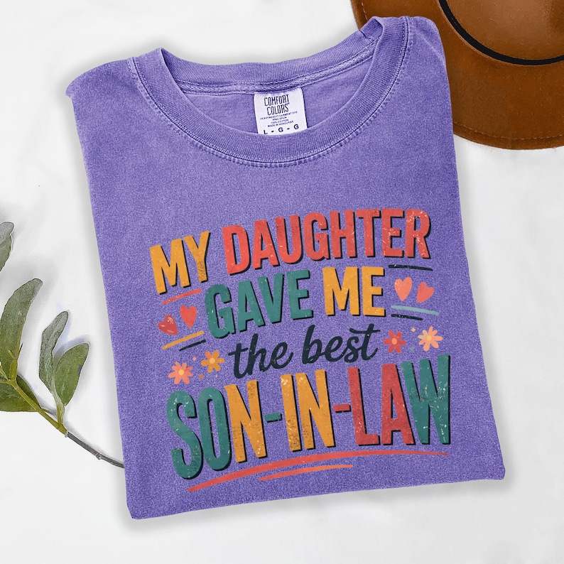 May include: A purple Comfort Colors t-shirt with the text "MY DAUGHTER GAVE ME the best SON-IN-LAW" in colourful lettering. The shirt has a crew neck and short sleeves. A brown hat is partially visible in the background.