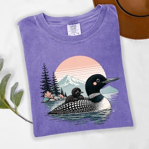 以下が含まれることがあります： 水面にいるアビとヒナのグラフィックが特徴の紫色のComfort Colors Tシャツ。背景には山と夕焼けが描かれています。このTシャツはクルーネックと半袖です。