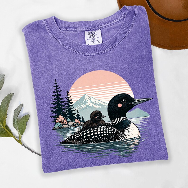 以下が含まれることがあります： 水面にいるアビとヒナのグラフィックが特徴の紫色のComfort Colors Tシャツ。背景には山と夕焼けが描かれています。このTシャツはクルーネックと半袖です。