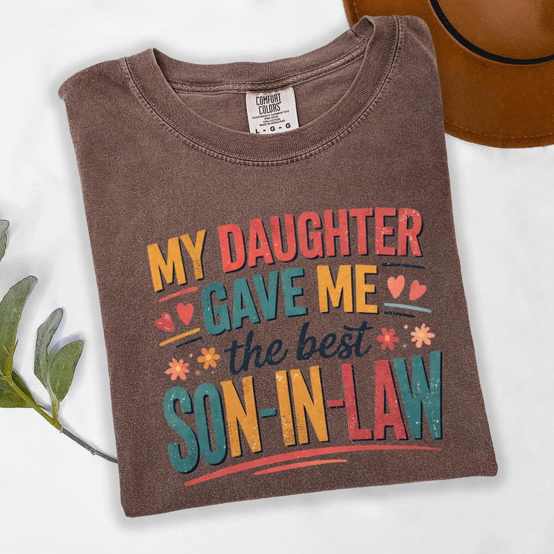 May include: Brown t-shirt with the text "MY DAUGHTER GAVE ME the best SON-IN-LAW" in colourful lettering. The shirt has a crew neck and a Comfort Colors tag. A brown hat is partially visible in the background.