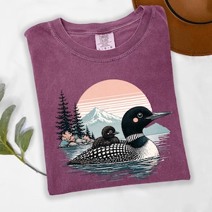 以下が含まれることがあります： 湖にいるハシグロアビとヒナのグラフィックが特徴の、マルーン色のComfort Colors Tシャツ。背景には山と夕焼けが描かれています。Tシャツはクルーネックと半袖です。Comfort Colorsのロゴが見えます。
