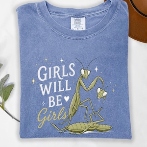 May include: A blue t-shirt with the words "GIRLS WILL BE Girls" and a graphic of praying mantises. The shirt is made of a soft material and has a crew neck. The Comfort Colors tag is visible.