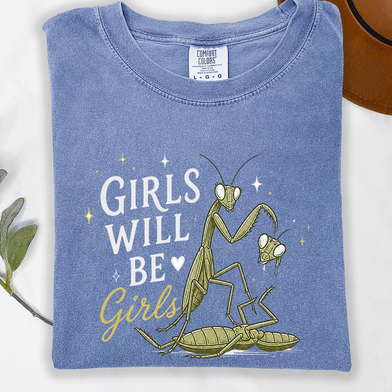 May include: A blue t-shirt with the words "GIRLS WILL BE Girls" and a graphic of praying mantises. The shirt is made of a soft material and has a crew neck. The Comfort Colors tag is visible.