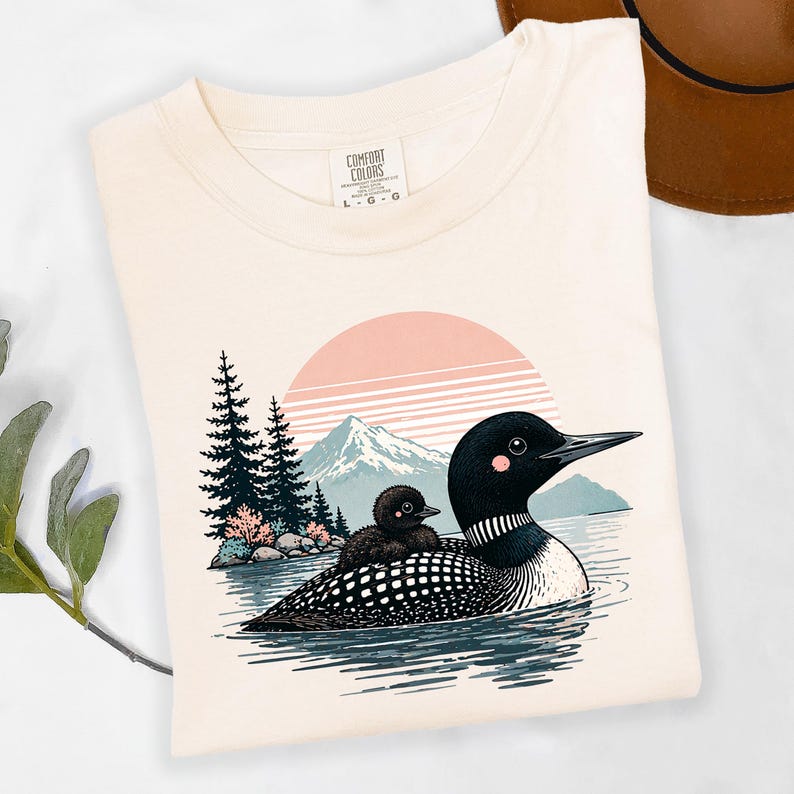 以下が含まれることがあります： クリーム色のTシャツ。湖にいるハシグロアビとヒナのグラフィック、ピンク色の夕日、山、木々が背景に描かれています。シャツにはComfort Colorsのロゴが入っています。