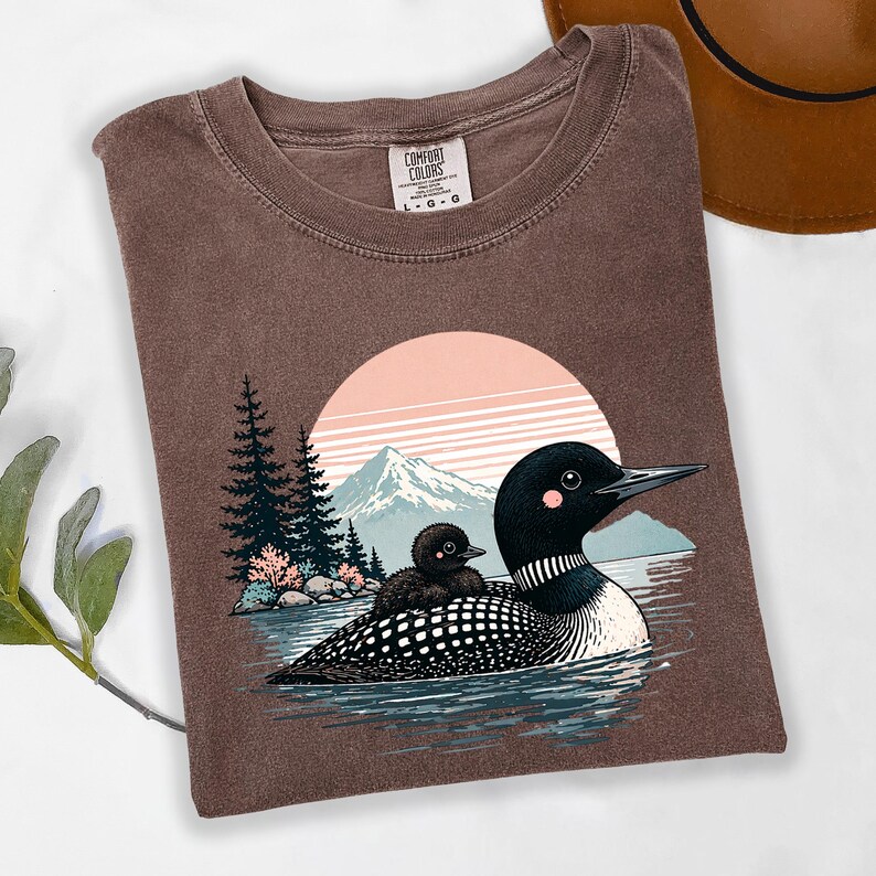 以下が含まれることがあります： 茶色のComfort Colors Tシャツ。湖にいるアビとヒナのグラフィック、山と夕日の背景が特徴です。Tシャツはクルーネックで、サイズはL-Gです。