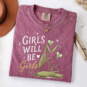 May include: A maroon t-shirt with the text "GIRLS WILL BE Girls" and a graphic of praying mantises. The shirt has a crew neck and short sleeves. A silver necklace with a pendant is draped across the shirt.