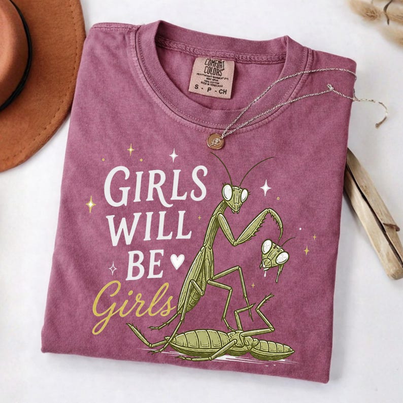 May include: A maroon t-shirt with the text "GIRLS WILL BE Girls" and a graphic of praying mantises. The shirt has a crew neck and short sleeves. A silver necklace with a pendant is draped across the shirt.