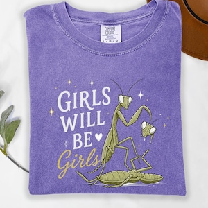 May include: A purple t-shirt with the text "GIRLS WILL BE Girls" in white and gold script. The shirt features a green praying mantis illustration with a whimsical design.