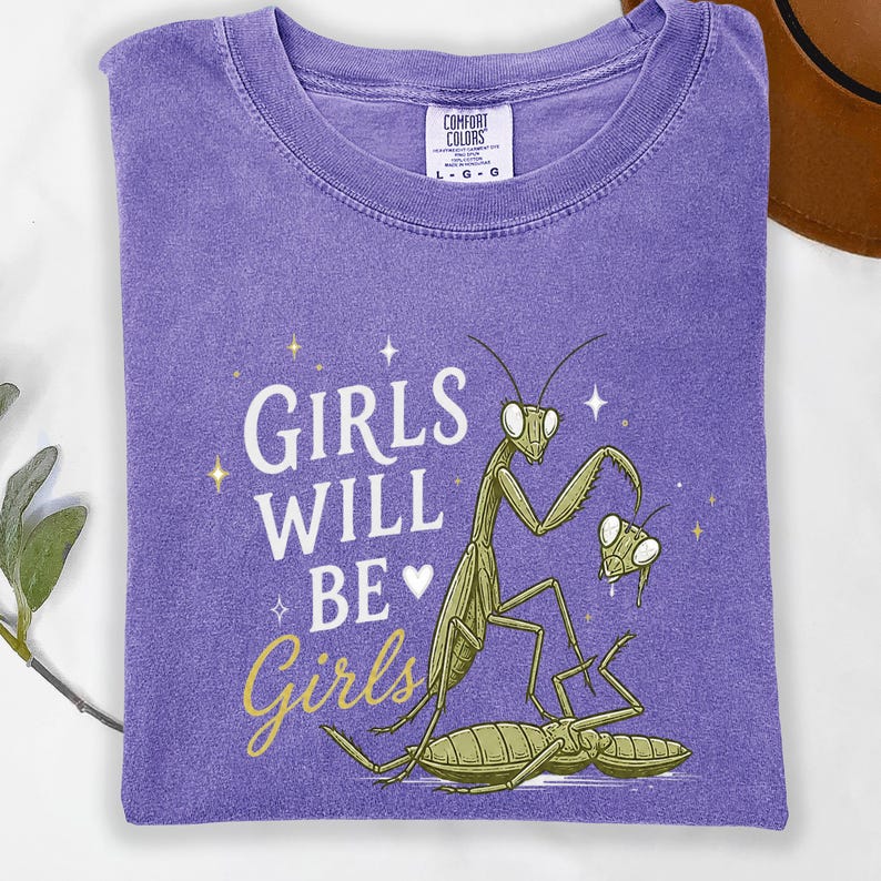 May include: A purple t-shirt with the text "GIRLS WILL BE Girls" in white and gold script. The shirt features a green praying mantis illustration with a whimsical design.