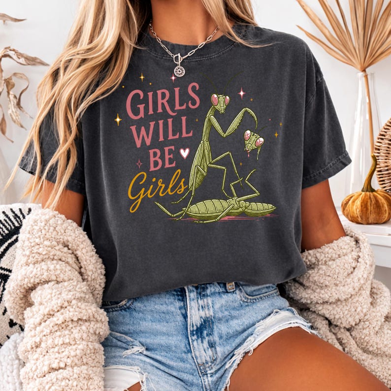 May include: Dark gray t-shirt with the text "GIRLS WILL BE GIRLS" in pink and yellow. The graphic features a green praying mantis illustration. The shirt is a casual style, suitable for everyday wear.