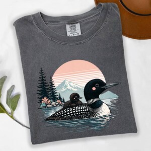 以下が含まれることがあります： 湖にいるアビとヒナのグラフィックが特徴のダークグレーのComfort Colors Tシャツ。ピンクとオレンジの夕日、山、木々が背景に描かれています。Tシャツはクルーネックと半袖です。