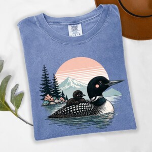 以下が含まれることがあります： 青色のComfort Colors Tシャツ。湖にいるハシグロアビとヒナのグラフィック、山と夕日の背景。クルーネックで、Comfort Colorsのタグが見えます。