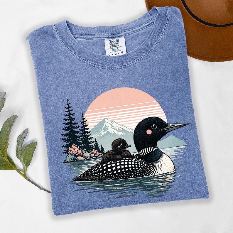 以下が含まれることがあります： 青色のComfort Colors Tシャツ。湖にいるハシグロアビとヒナのグラフィック、山と夕日の背景。クルーネックで、Comfort Colorsのタグが見えます。
