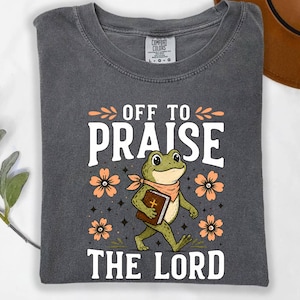 May include: A charcoal gray t-shirt featuring a graphic of a green frog wearing a neckerchief and carrying a brown book. The text reads "OFF TO PRAISE THE LORD" in white letters, surrounded by orange flowers and small black star accents.