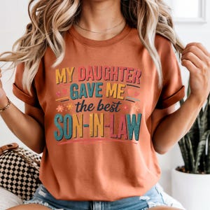 May include: A rust-coloured t-shirt featuring the text "MY DAUGHTER GAVE ME the best SON-IN-LAW" in a layered, colourful design. The shirt has rolled-up sleeves and a crew neck. The text uses a mix of fonts and colours.