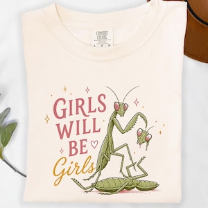 May include: Cream-colored t-shirt featuring a graphic of a praying mantis with the text "Girls Will Be Girls" in pink and gold. The design includes a heart and star accents, and the shirt has a Comfort Colors tag.