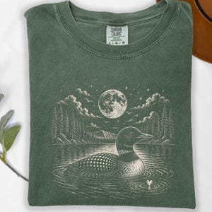 May include: Green Comfort Colors t-shirt featuring a detailed graphic of a loon on a lake under a full moon and starry sky. The design includes trees and a heart. The shirt has a classic crew neck and the Comfort Colors tag is visible.