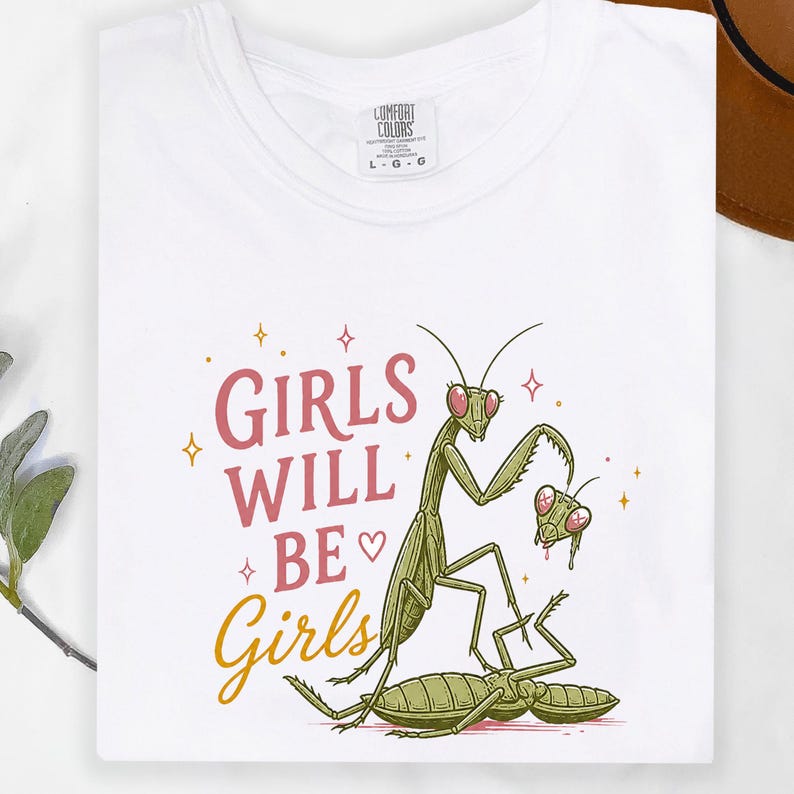 May include: White t-shirt with a graphic of two green praying mantises and the text "GIRLS WILL BE Girls" in pink and gold. The shirt is made of a soft, comfortable material.