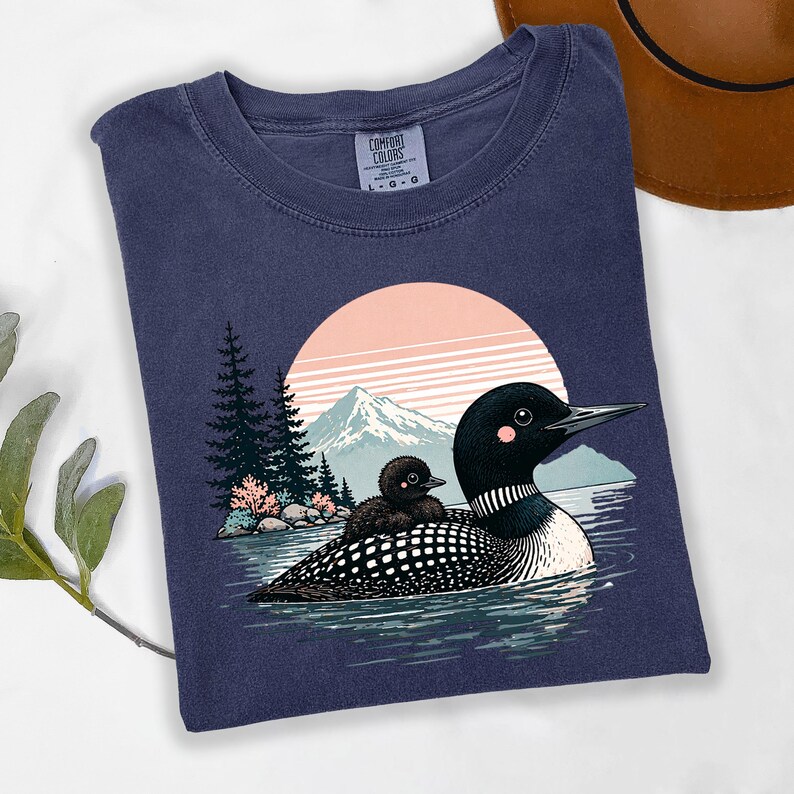 以下が含まれることがあります： ネイビーブルーのComfort Colors Tシャツ。水面にいるハシグロアビとヒナのグラフィック。デザインにはピンクと白の夕日、山、木が含まれています。シャツのサイズはL-Gです。