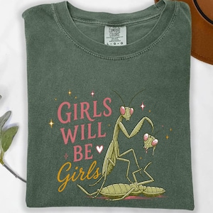 May include: Green t-shirt with the text "GIRLS WILL BE Girls" in pink and yellow. The shirt features a cartoon illustration of praying mantises. The shirt is a Comfort Colors brand.