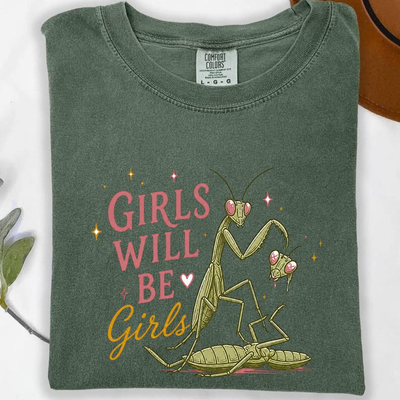 May include: Green t-shirt with the text "GIRLS WILL BE Girls" in pink and yellow. The shirt features a cartoon illustration of praying mantises. The shirt is a Comfort Colors brand.