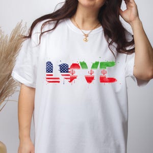 May include: White t-shirt with the word "LOVE" in a watercolor design. The "L" and "E" are in the colors of the Iranian flag, while the "O" and "V" are in the colors of the American flag. The shirt is a casual style.