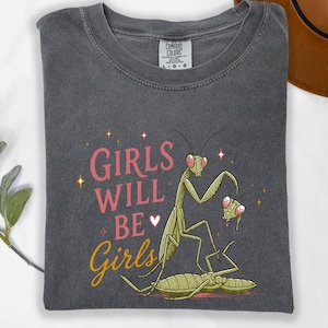 May include: Dark gray t-shirt with the text "GIRLS WILL BE Girls" in pink and yellow. The shirt features a cartoon illustration of praying mantises. The shirt is made of a soft material.