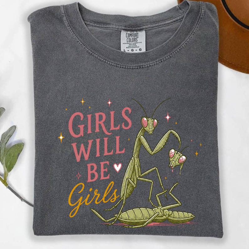 May include: Dark gray t-shirt with the text "GIRLS WILL BE Girls" in pink and yellow. The shirt features a cartoon illustration of praying mantises. The shirt is made of a soft material.