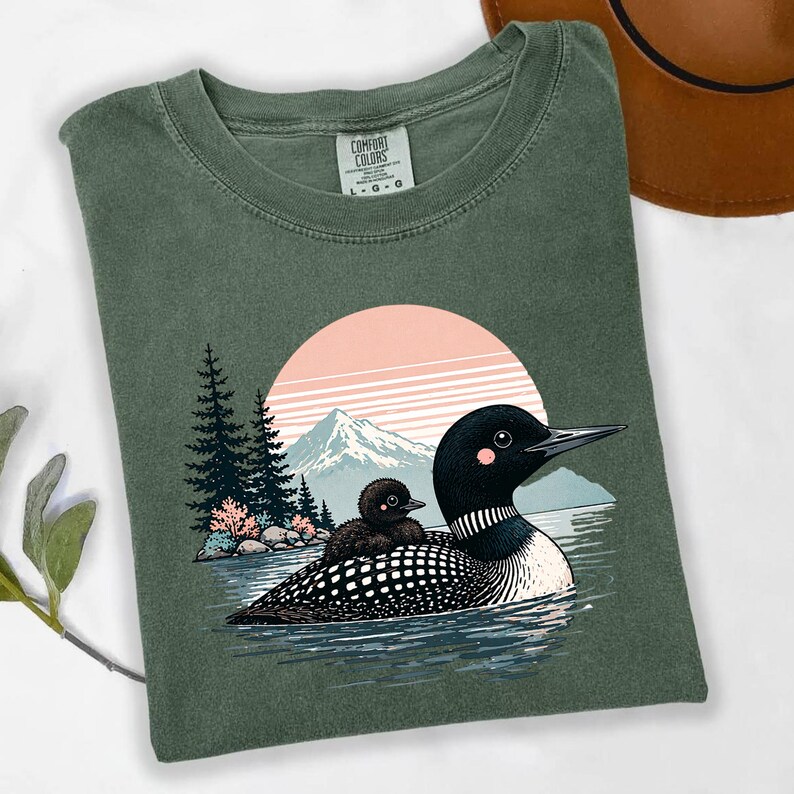 以下が含まれることがあります： 緑色のComfort Colors Tシャツ。オオハシとヒナが水面にいるグラフィックで、背景には山と夕焼けが描かれています。デザインには木と水が含まれており、自然をテーマにした美学を作り出しています。