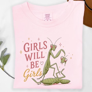 May include: A light pink t-shirt with the words "GIRLS WILL BE Girls" in a mix of pink and gold script. The shirt features a cartoon illustration of praying mantises in green and pink.