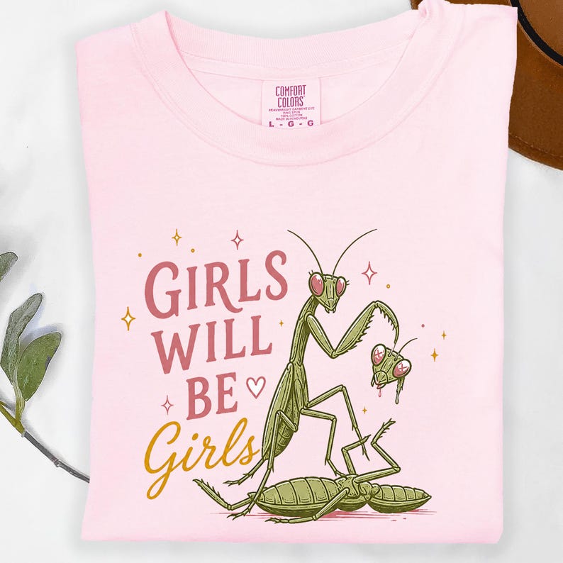 May include: A light pink t-shirt with the words "GIRLS WILL BE Girls" in a mix of pink and gold script. The shirt features a cartoon illustration of praying mantises in green and pink.