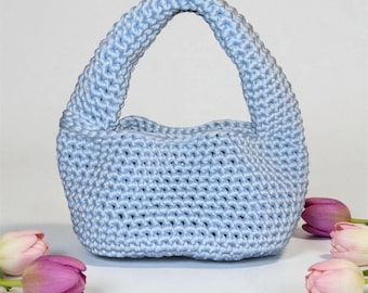 May include: A pale blue crochet handbag with a curved handle. The bag is surrounded by pink and white tulips, creating a soft, feminine aesthetic. The bag's texture is visible, showing the crochet stitches.