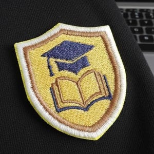 May include: Embroidered shield-shaped patch with a graduation cap and open book design. The patch has a yellow background, brown and blue details, and a white border. It is attached to black fabric.