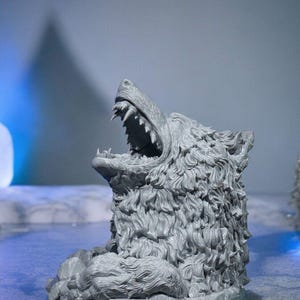 May include: A gray, detailed sculpture of a wolf's head with its mouth open, revealing sharp teeth. The sculpture has a textured, fur-like appearance and sits on a rocky base. The background is a soft blue and gray.