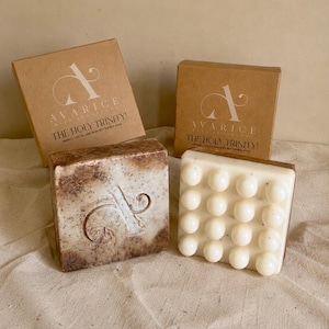 May include: Two square bar soaps and their matching brown cardboard boxes labeled Avarice The Holy Trinity. One soap is mottled brown and white with an embossed logo, while the other is solid white with a grid of sixteen raised circular bumps.